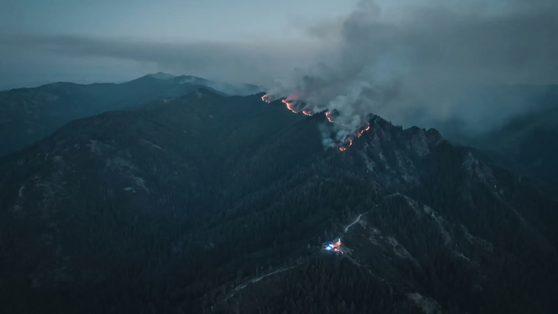 A fire line burning along a forested mountain ridge at night, with the lights of staged apparatus visible on a road below.