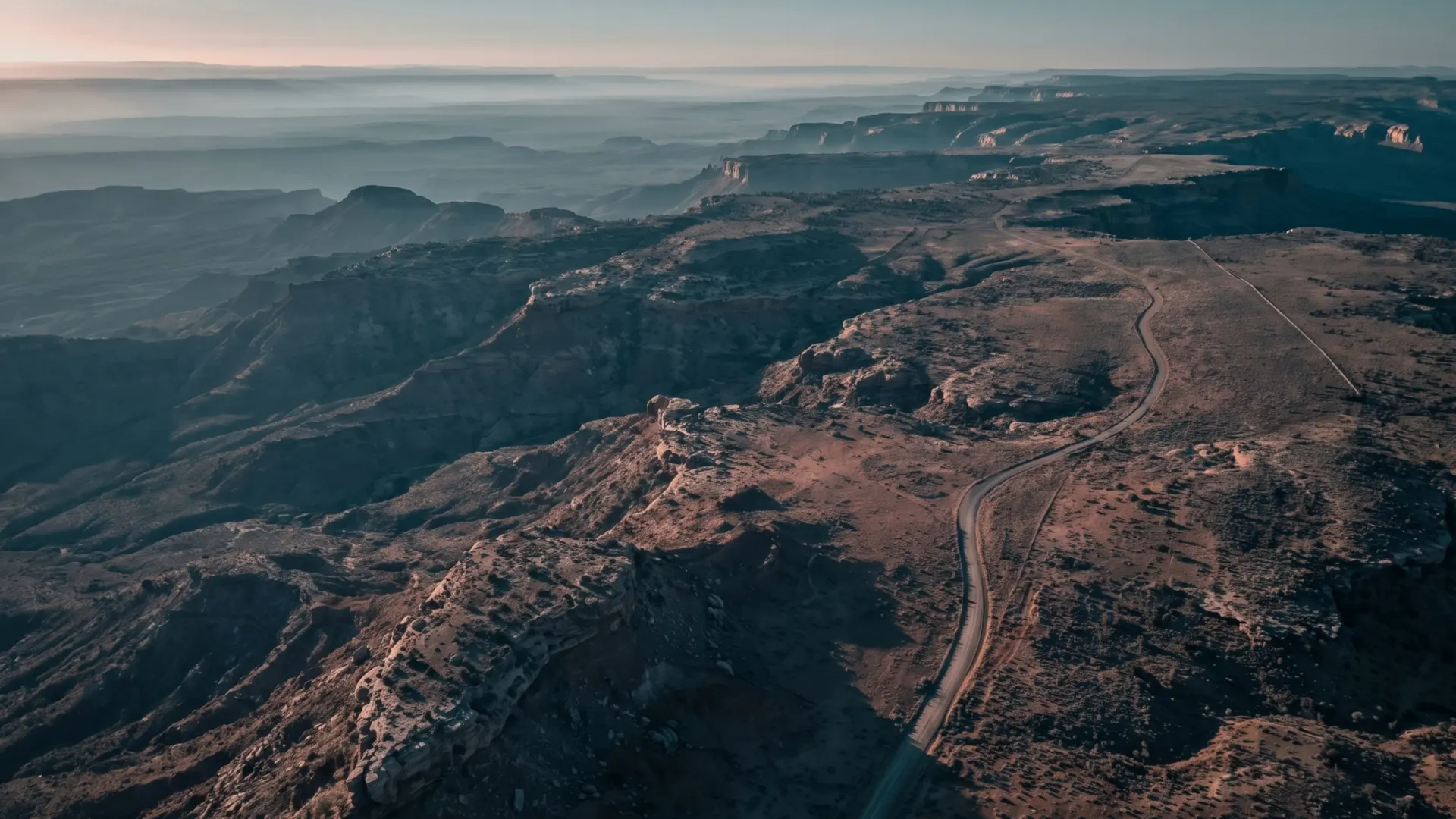 A desert mesa at first light, a two-track road running along the rim and a distant fence line on the plateau beyond.